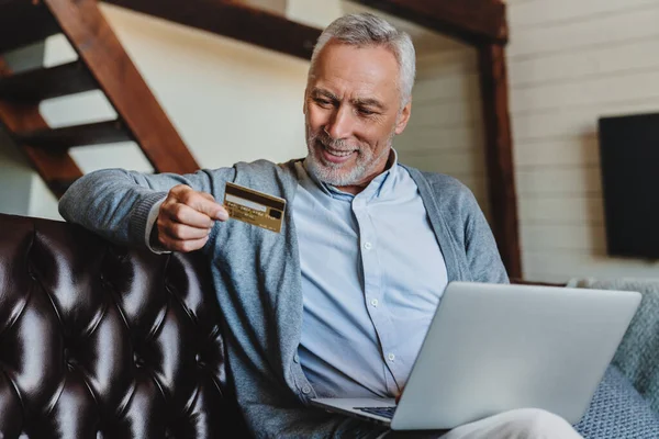 Szczęśliwy Starszy Człowiek Pomocą Laptopa Trzymając Kartę Kredytową Podczas Siedzenia — Zdjęcie stockowe