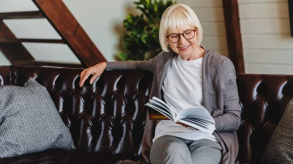 Ηλικιωμένη Γυναίκα Γυαλιά Κάθεται Στον Καναπέ Και Διαβάζει Ενδιαφέρον Βιβλίο — Φωτογραφία Αρχείου