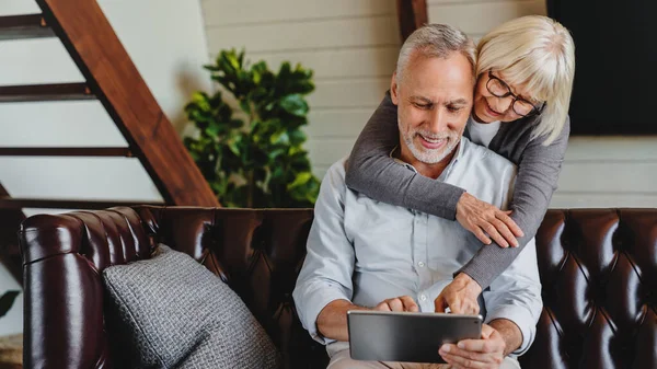 Oudere Paar Glimlachen Kijken Naar Digitale Tablet Geknuffeld Woonkamer Rechtenvrije Stockfoto's