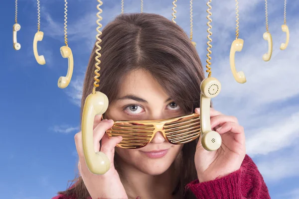 flirting woman adjusting golden party glasses and standing against blue sky with hanging old fashioned phones