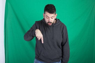 Kapüşonlu ve yeşil arka planı poz, aşağı bakıyor ve parmak aşağı dönük adam 