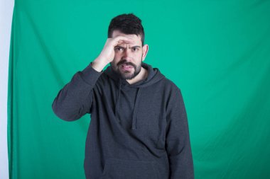 Kapüşonlu ve yeşil arka planı poz adam başında elini tutarak ve arama sırasında kameraya bakan 