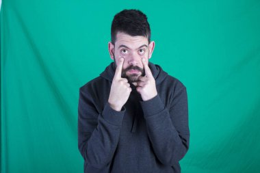 Kapüşonlu ve yeşil arka planı poz, kameraya bakarak ve parmaklarını gözlerine tutan adam