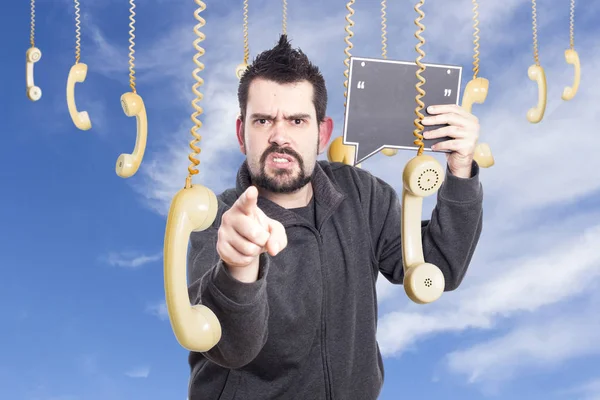 angry man against sky surrounded by hanging old fashioned phones handsets, pointing finger on camera and  holding speech bubble boar