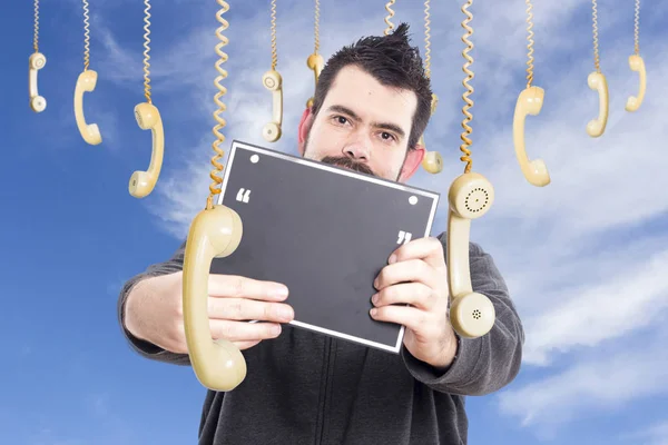 man against sky surrounded by hanging old fashioned phones handsets, holding speech bubble board