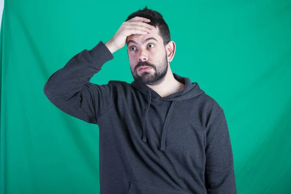 shocked man wearing hoodie and posing against green background, holding hand at head