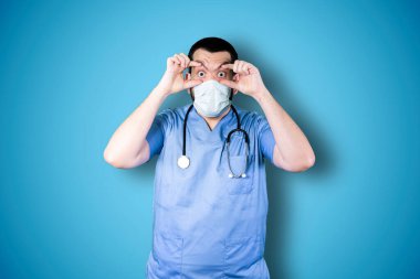 Maskeli doktor mavi arka plan stüdyosunun üzerinde şaşırmış yüz ifadesiyle