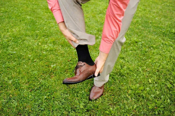 The man takes off his shoe, standing on the grass. Classic suit, black socks.