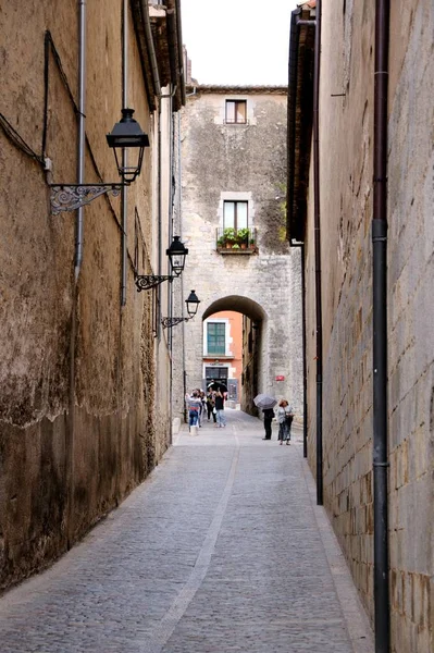 Medieval street of Girona. A narrow street with stone walls, street lights and an archway. Tourists are walking along the street.