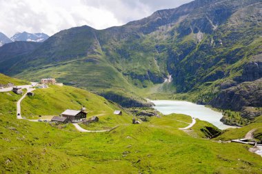 A blue mountain lake between the green slopes of the mountains. Several small houses and hotel buildings. Beautiful curving mountain road, Grossglockner Hochalpenstrasse, Austria. Summer day. clipart