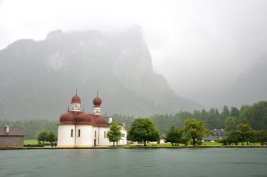 St Bartholomew's kilise Konigssee. Su Dağları nın arka planı göster. Bulutlu yağmurlu bir gün, sis, çamurlu karanlık su göle. Bavyera, Almanya.