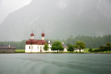 St Bartholomew's kilise Konigssee. Su Dağları nın arka planı göster. Bulutlu yağmurlu bir gün, sis, çamurlu karanlık su göle. Bavyera, Almanya.