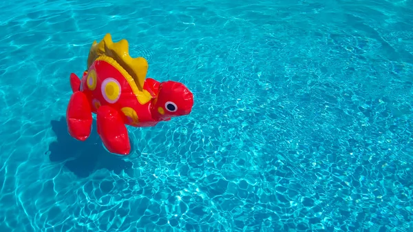 An inflatable red dinosaur in clear rippling pool water. Funny baby toy floats isolated in blue water. Summer vacation sale concept. Copy space for text