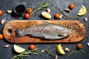 Ahşap doğrama tahtası üzerinde ham alabalık. Malzemeler biberiye, limon, domates, sarımsak, tuz, karabiber. Siyah arka plan, üstten görünüm