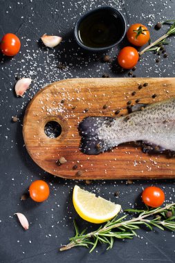 Ahşap doğrama tahtası üzerinde ham alabalık. Malzemeler biberiye, limon, domates, sarımsak, tuz, karabiber. Siyah arka plan, üstten görünüm