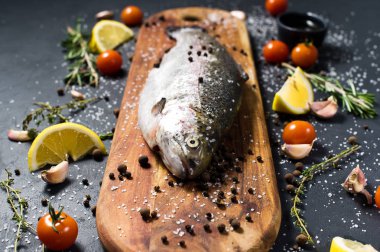 Ahşap doğrama tahtası üzerinde ham alabalık. Malzemeler biberiye, limon, domates, sarımsak, tuz, karabiber. Siyah arka plan, yan görünüm