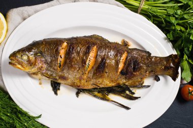 Beyaz bir levha, dal dereotu ve maydanoz üzerinde Izgara Alabalık. Siyah arka plan, üstten görünüm