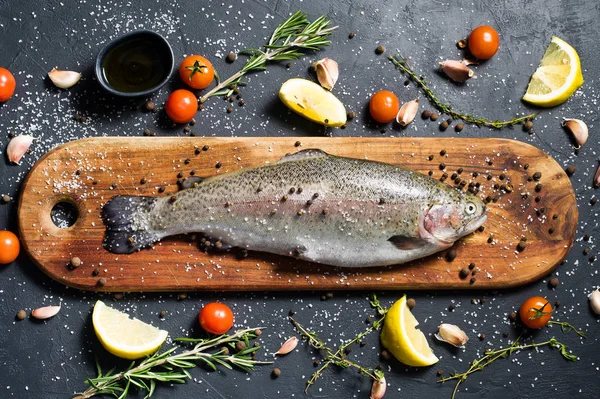 Ahşap doğrama tahtası üzerinde ham alabalık. Malzemeler biberiye, limon, domates, sarımsak, tuz, karabiber. Siyah arka plan, üstten görünüm