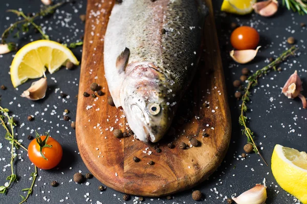 Ahşap doğrama tahtası üzerinde ham alabalık. Malzemeler biberiye, limon, domates, sarımsak, tuz, karabiber. Siyah arka plan, yan görünüm