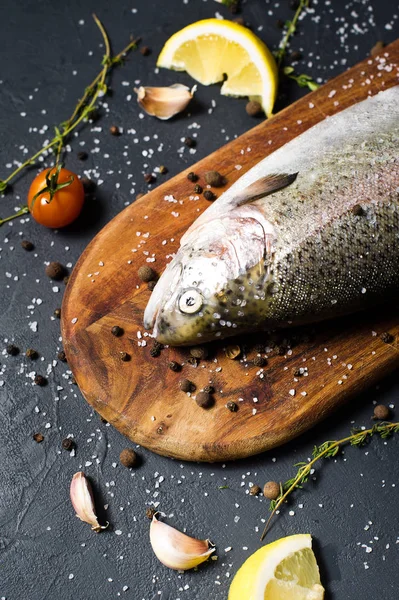 Ahşap doğrama tahtası üzerinde ham alabalık. Malzemeler biberiye, limon, domates, sarımsak, tuz, karabiber. Siyah arka plan, yan görünüm
