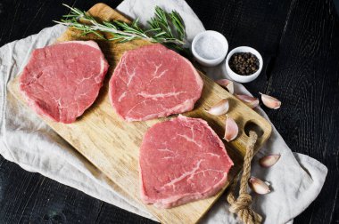 Üç ham sığır eti biftek Ahşap doğrama tahtası, sarımsak ve biberiye bir delikanlı. Üstten Görünüm, siyah arka plan