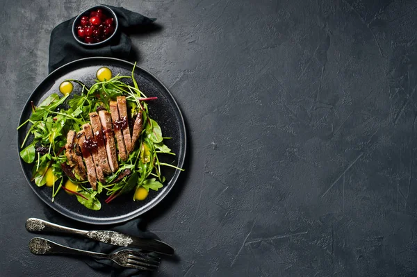 Salad with beef tenderloin steak, arugula and chard on a black plate. Cranberry sauce. Top view, black background, space for text