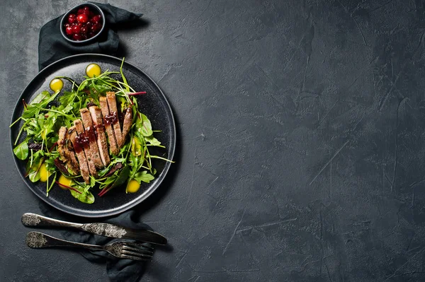 Salad with beef steak, arugula and chard on a black plate. Cranberry sauce. Top view, black background, space for text
