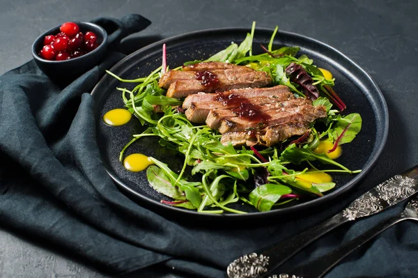 Salad with beef tenderloin steak, arugula and chard on a black plate. Cranberry sauce. Top view, black background, close up