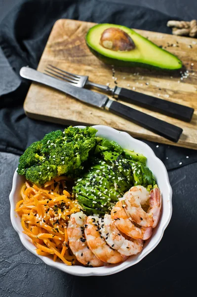 The Buddha bowl a balanced healthy meal. Ingredients: shrimps, avacado, carrots, broccoli and rice. Black background, side view