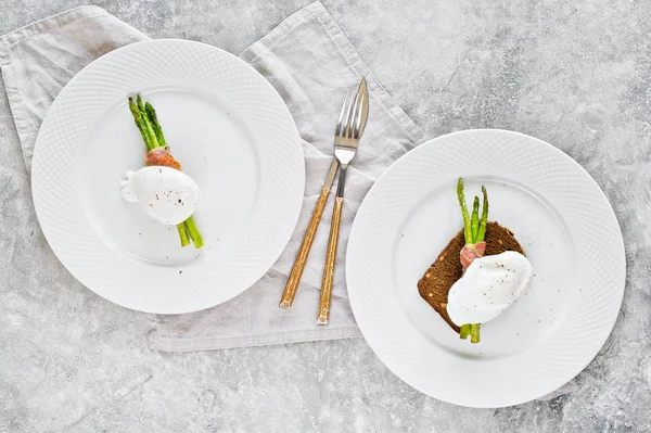 Deux assiettes avec un œuf poché sur des asperges rôties enveloppées dans du bacon. Fond gris, vue de dessus . — Photo