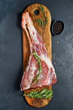 Tahta bir kesme tahtasında keçi ham bacak. Biberiye, keklik, karabiber. Siyah arka plan, üst görünüm, metin alanı