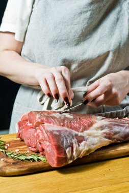 Şef, ahşap bir doğrama tahtasında çiğ kuzu bacağı pişiriyor. Biberiye, keklik, karabiber. Siyah arka plan, yan görünüm, metin alanı
