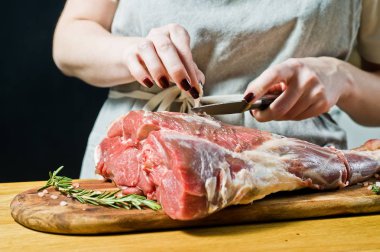 Şef, ahşap bir doğrama tahtasında çiğ keçi bacağı pişiriyor. Biberiye, keklik, karabiber. Siyah arka plan, yan görünüm, metin alanı