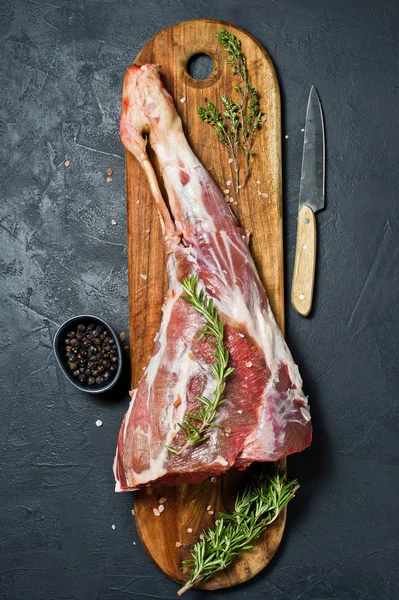 Ahşap bir tahta doğrama üzerinde kuzu ham bacak. Biberiye, keklik, karabiber. Siyah arka plan, üst görünüm