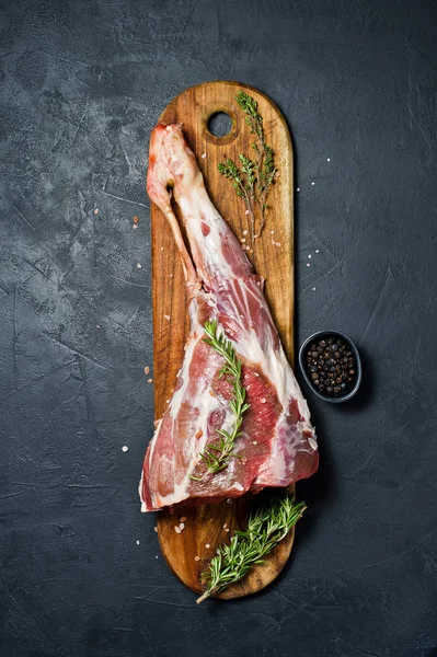 Ahşap bir tahta doğrama üzerinde kuzu ham bacak. Biberiye, keklik, karabiber. Siyah arka plan, üst görünüm