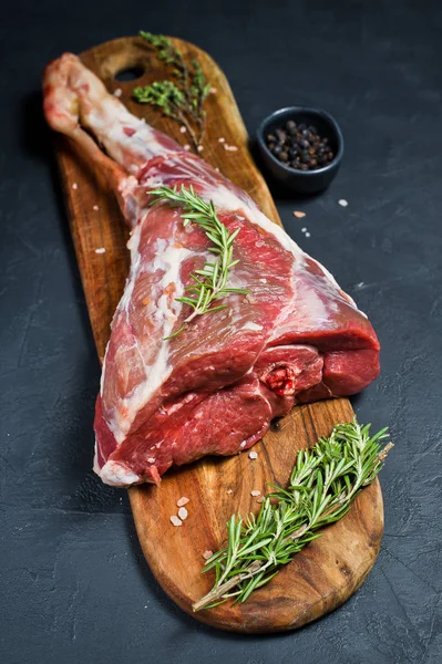 Ahşap bir tahta doğrama üzerinde kuzu çiğ bacak. Biberiye, keklik, karabiber. Siyah arka plan, yan görünüm