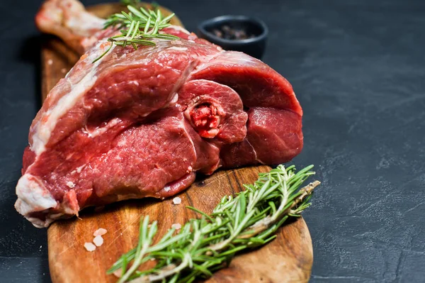 Ahşap bir tahta doğrama üzerinde kuzu çiğ bacak. Biberiye, keklik, karabiber. Siyah arka plan, yan görünüm, metin alanı