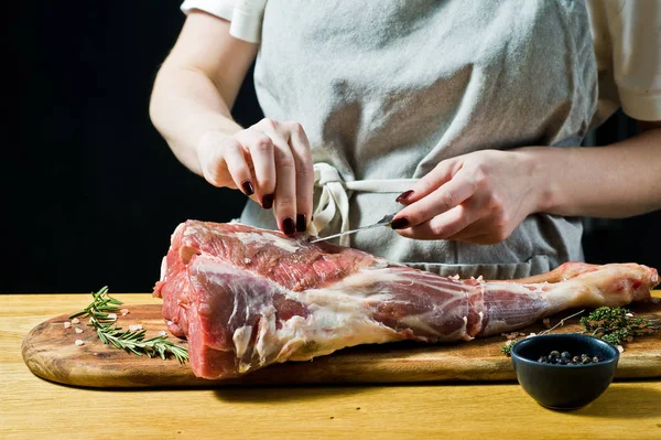 Şef, ahşap bir doğrama tahtasında kuzu çiğ bir bacak keser. Biberiye, keklik, karabiber. Siyah arka plan, yan görünüm, metin alanı