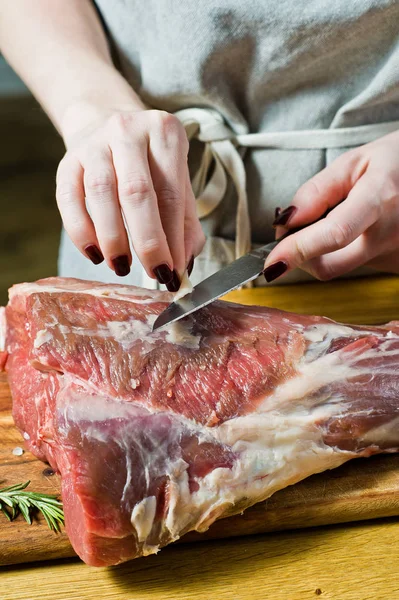 Şef, ahşap bir doğrama tahtasında kuzu çiğ bir bacak keser. Biberiye, keklik, karabiber. Siyah arka plan, üst görünüm, metin alanı