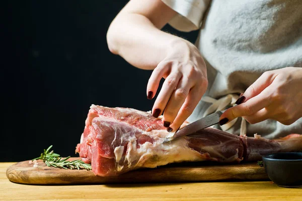 Şef, ahşap bir doğrama tahtasında çiğ keçi bacağını keser. Biberiye, keklik, karabiber. Siyah arka plan, yan görünüm, metin alanı