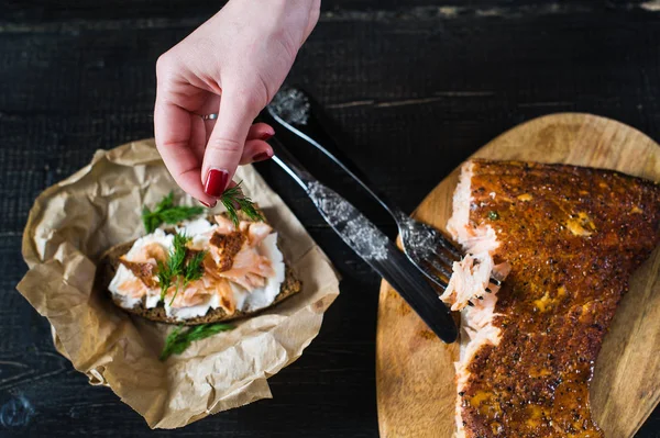 El chef espolvorea perejil sobre un sándwich con filete de salmón ahumado sobre pan negro y queso suave — Foto de Stock