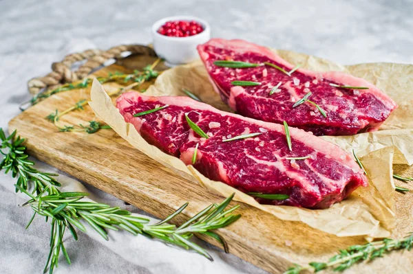 Syrové hovězí bifteky Angus na dřevěné sekací desce s rozmarýnem a růžovým pepřem. Šedé pozadí, pohled shora. — Stock fotografie