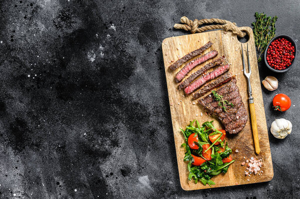 Grilled sliced Denver steak on a cutting board. BBQ beef. Black background. Top view. Copy space.