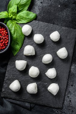Üzerinde fesleğen yaprağı ve pembe biber olan taş bir tahtada mini mozzarella peyniri topları. Siyah arka plan. Üst görünüm.