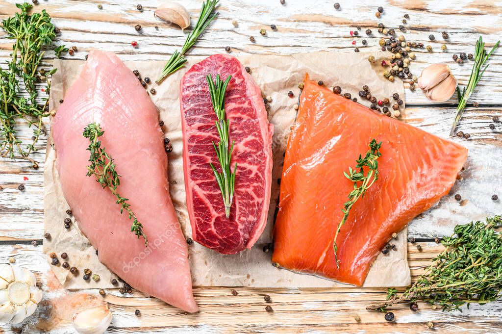 Different types of steaks on wooden table with herbs. Beef top blade, salmon fillet and Turkey breast. Organic fish, poultry and beef meat. White background. Top view.