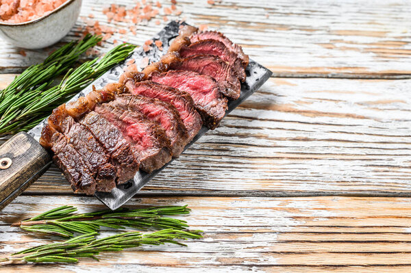 Grilled top sirloin cap or picanha steak on a meat cleaver with herbs. White wooden background. Top view. Copy space.