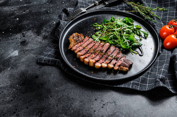 Cooked grilled strip loin steak, marbled beef meat with arugula. Black background. Top view. Copy space