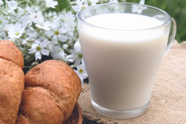 Kruvasan ve beyaz çiçeklerin yanında bir bardak süt var..