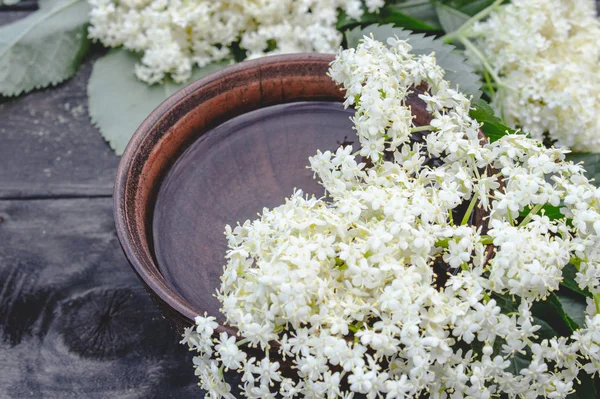 White flowers and leaves of elderberry lie on a bowl on a rustic wooden background.