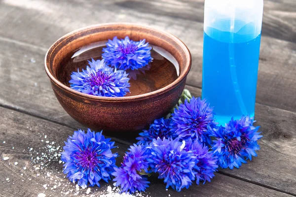 青いトウモロコシの花は、古いボードの背景に対してコーンフラワー水のボトルの近くのボウルの中に横たわっています. — ストック写真